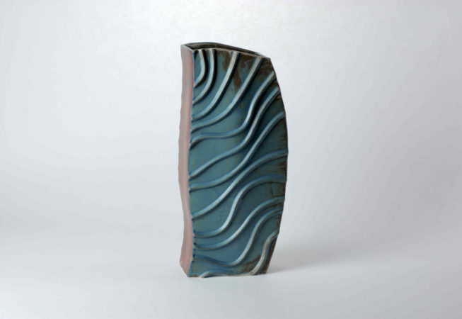 Eine hohe geschwungene aufgebaute Vase. Das Einzelstück hat zwei braune Seiten und zwei blaue Seiten. Die blauen Seiten haben aufgelegte Tonwülste, die an fließendes Wasser erinnern sollen.